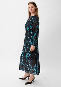 Schwarzes Kleid mit langen Ärmeln und einem Ausschnitt in Form eines Schlüssellochs. Das Kleid hat ein blaues abstraktes Muster und eine fließende Silhouette, kombiniert mit schwarzen Absätzen.