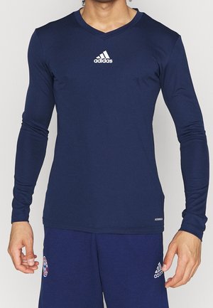 Mand iført mørkeblå Adidas langærmet V-hals trøje og matchende shorts med Adidas- og Bayern München-logoer.