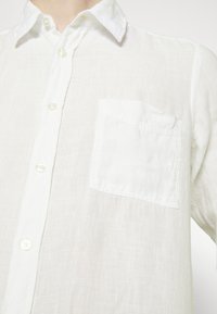 Camisa de lino blanca con cuello abotonado, un bolsillo en el pecho y botones perlados, que presenta una tela texturizada y transpirable.