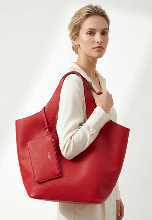 Vrouw in wit blouse draagt grote gestructureerde rode leren tote bag met bijpassend ritsvakje over de schouder.
