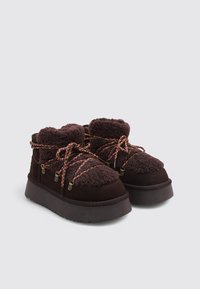 Botas de ante marrón con parte superior de forro polar texturizado, suelas gruesas de goma y cordones trenzados multicolores. Presentan detalles con doble cremallera.