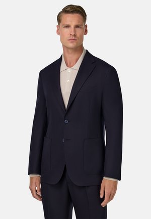 Blazer blu navy con due tasche frontali, due bottoni e rever a punta. Sotto, una polo beige con tre bottoni e colletto.