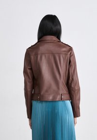 Jachetă de piele maro, stil biker, cu epoleți pe umeri, design ajustat și accente cu butoane, asortată cu o fustă plisată de culoare teal.