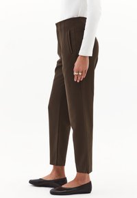 Donna che indossa pantaloni slim-fit a vita alta marrone scuro, una maglietta bianca a maniche lunghe, ballerine nere e anelli d'oro sulla mano, in piedi di lato.