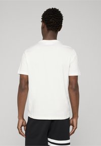 Hvid bomuldst-shirt med rund hals, korte ærmer og en afslappet pasform, set bagfra, kombineret med sorte shorts med hvide striber.