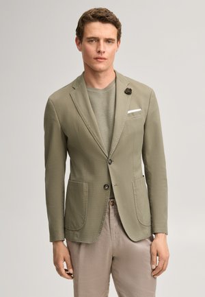 Mann trägt hellolivfarbenen Blazer mit zwei Knöpfen, Seitentaschen, einer floral gestalteten Reversnadel, grauem Strickpullover und beigen Hosen vor einfachem Hintergrund.