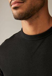 Chemise noire côtelée à manches courtes avec un col rond, présentant un tissu texturé et une coupe ajustée. Vue rapprochée du col et de l'épaule.