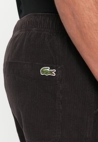 Corduroy pants in dark brown with vertical ridges. Features a small embroidered green crocodile logo on the back pocket.