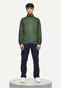 Zielona bluza z zamkiem na wysokości szyi, noszona z ciemnymi dżinsami i białymi trampkami, wyeksponowana na okrągłym betonowym piedestale.