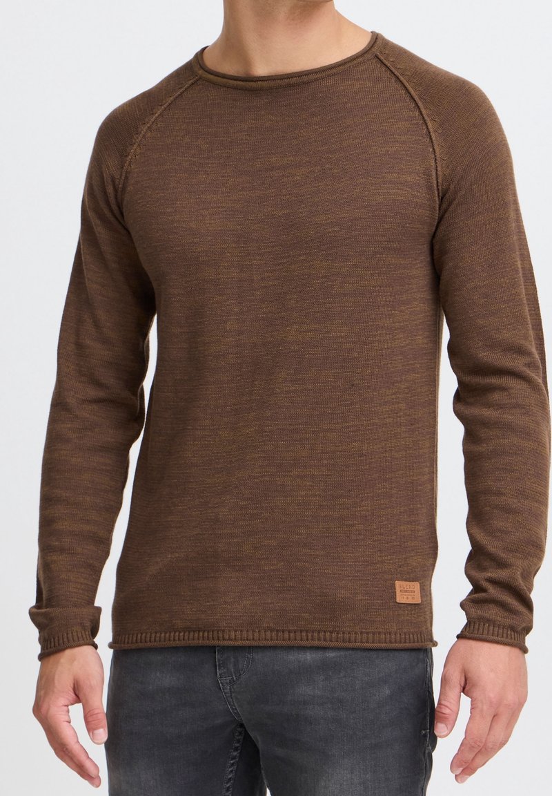 Homme portant un pull en maille marron à manches longues et un jean gris foncé sur un fond uni.