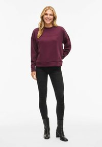 Donna in piedi e sorridente, che indossa una felpa viola con il testo "MINIMALISM", jeans skinny neri e stivaletti neri con tacco su sfondo bianco.