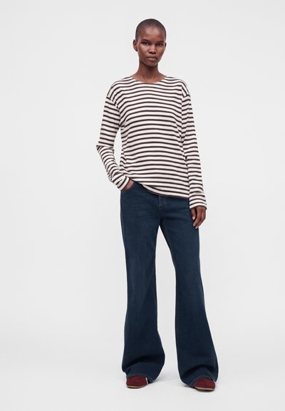 Femme debout, une main dans la poche, portant une chemise à manches longues blanche et marron à rayures et un jean large foncé avec des chaussures rouges.
