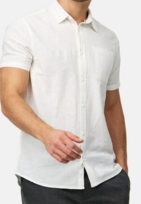 Mężczyzna w białej lnianej koszuli z krótkim rękawem, zapinanej na guziki, z przednią kieszonką na piersi, w połączeniu z ciemnoszarymi spodniami, z ręką częściowo wyciągniętą.