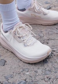 Scarpe sportive rosa chiaro con parte superiore texturizzata, chiusura con lacci e suola ammortizzata. Indossate con calzini bianchi alla caviglia su una superficie di ghiaia.