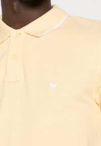 Gul polo skjorta med texturerad tyg, tre knappar vid kragen och en liten vit logotyp på vänster sida.