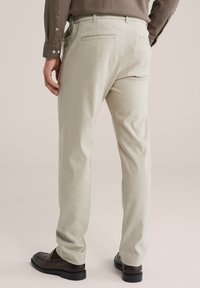 Homme portant un pantalon beige, une chemise marron et des chaussures en cuir marron foncé, debout devant un fond clair uni.