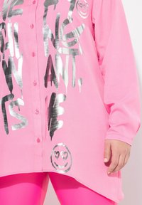 Pink Langarmshirt mit silbernem Grafiktext und einem lächelnden Emoji am Saum. Weicher Stoff mit lockerem Schnitt. Knopfverschluss.