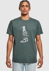 Sötétzöld pamut póló, amelyen egy grafika látható, ami egy sneakerben álló sirályt ábrázol, részletes textúrákkal mind a madáron, mind a lábbelin.