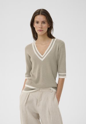 Frau trägt einen beigen, gerippten V-Ausschnittpullover mit weißen Streifen und hellbeige, plissierte Hose, steht mit den Händen in den Taschen.