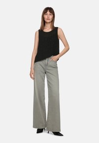 Schwarzes ärmelloses Oberteil mit strukturiertem Design, kombiniert mit hellgrauen weit geschnittenen Jeans und schwarzen spitzen Schuhen, stehend mit einer Hand in der Hüfte.