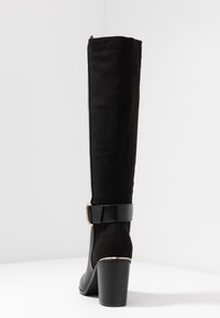 Black over-the-knee boot made of suede with a glossy black leather strap and gold-tone buckle. Features a chunky black heel.