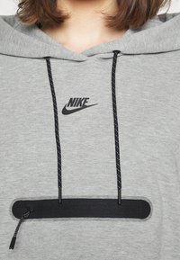 Sudadera gris hecha de material suave, con un logo negro de Nike, cordones negros con un detalle texturizado y un bolsillo con cremallera horizontal.