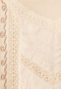 Tissu crème avec broderie marron verticale à gauche et bandes de dentelle diagonales formant un motif chevron.