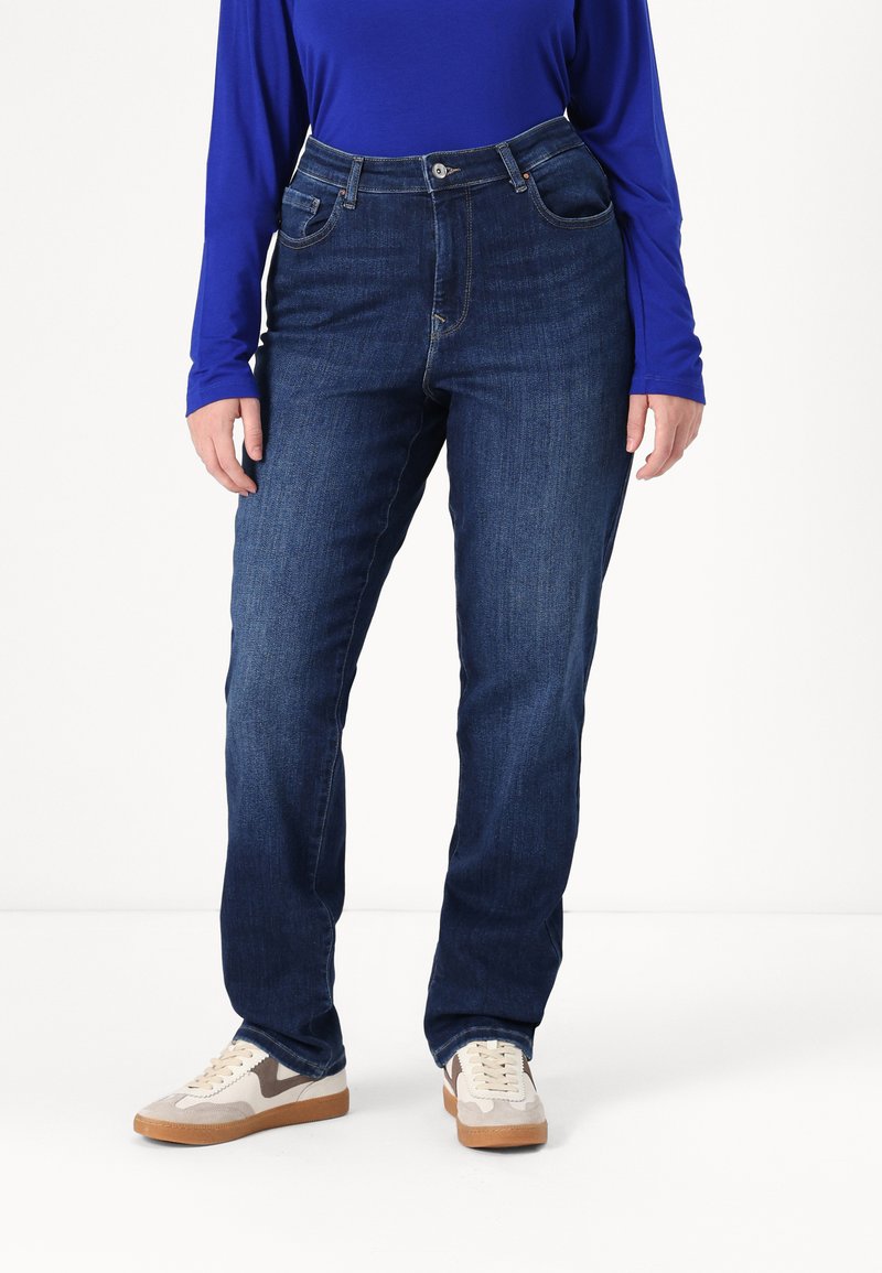 Jean en denim bleu foncé à taille haute, avec un design cinq poches et une texture lisse. Associé à des baskets beiges avec un accent à motifs.