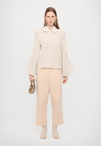 Bluză texturată bej deschis cu guler ascuțit, mâneci lejere tip balon; asortată cu pantaloni deschiși la culoare și o geantă cu model. Șosete deschise cu dantelă galbenă.