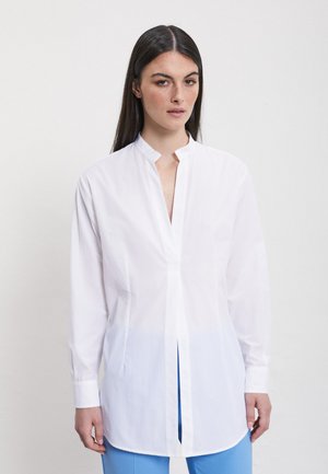 Giovane donna con lunghi capelli scuri che indossa una camicetta bianca a maniche lunghe con scollo a spacco e pantaloni azzurri su uno sfondo uniforme.
