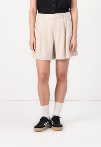 Shorts plissés beiges en tissu texturé, dotés de poches latérales, d'une taille haute et d'une coupe décontractée, associés à des baskets noires.