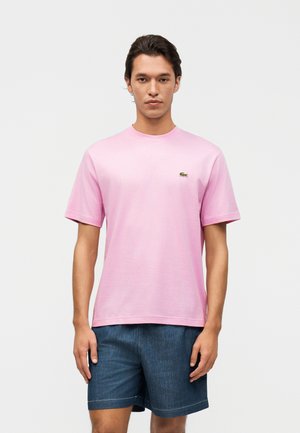 Homme portant une chemise à manches courtes rose clair avec un petit logo animalier et un short en jean bleu foncé, debout devant un fond uni.