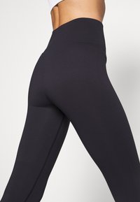 Czarne legginsy z wysokim stanem, wykonane z rozciągliwego materiału, charakteryzujące się gładką teksturą i detalami wzdłuż szwów.