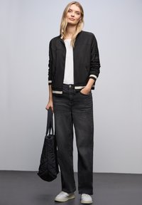 Veste bomber noire avec des poignets rayés, t-shirt blanc, jeans foncés à coupe ample, baskets blanches et sac matelassé noir.