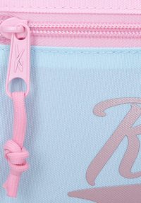 Primer plano de una cremallera rosa con un tirador de tela anudado en una bolsa azul claro que presenta una letra "K" estilizada parcial en rosa.
