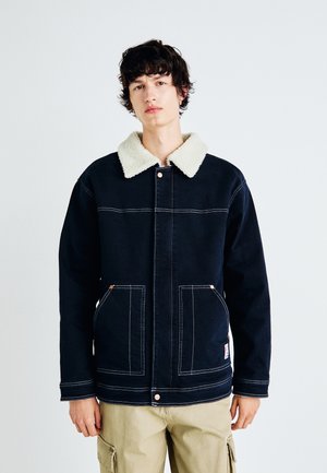 Giacca blu navy con colletto in sherpa crema, dotata di due tasche frontali, cuciture visibili e dettagli in ottone sui bottoni frontali.