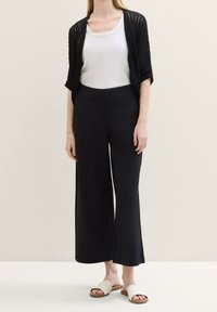 Schwarze weit geschnittene Hose mit glatter Textur, kombiniert mit einem weißen Tanktop und einer leichten schwarzen Strickjacke. Getragen mit weißen Sandalen.