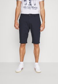 Shorts bleu marine avec un motif subtil, fabriqués en tissu léger. Portés avec un t-shirt blanc ajusté et des baskets blanches avec des accents colorés.