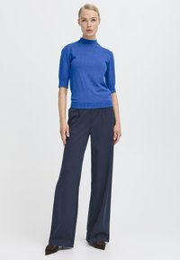 Niebieska krótka bluzka z wysokim dekoltem, zestawiona z ciemnymi spodniami typu wide-leg i brązowymi czółenkami. Gładka tkanina, elegancki krój.