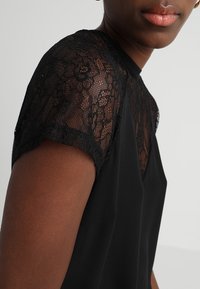 Black top featuring short sleeves made of intricate black lace. Smooth fabric below with a round neckline and fitted silhouette.