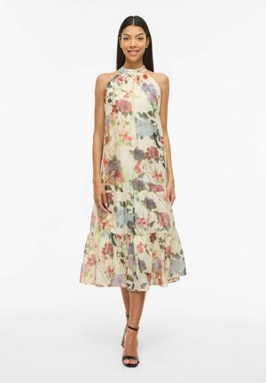 Žena oblečená v bezrukávových midi šatách s halterom a kvetinovým vzorom a čiernych podpätkoch s remienkami okolo členkov stojaca na bielom pozadí.