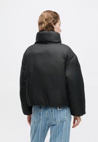 Čierna prešívaná bunda s vysokým golierom, s hladkým povrchom a nadrozmerným strihom. Nosená cez svetlo modré pruhované džínsy.