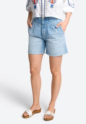Woman wearing light blue high-waist denim shorts, white embroidered blouse, and white slide sandals with wooden soles, hands in pockets.