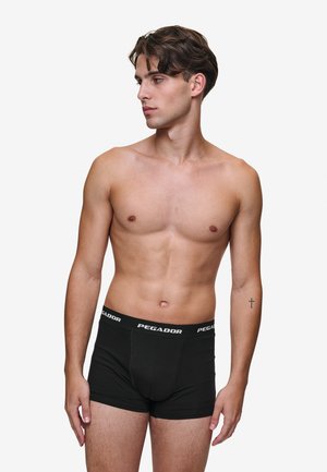 Boxer shorts noirs avec une texture douce, dotés d'une large ceinture élastique ornée du logo "PEGADOR" en blanc. Le modèle est torse nu, vu de côté.