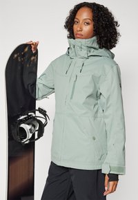 Svetlozelená nepremokavá bunda s vysokým golierom, zipsovými vreckami a nastaviteľnými lemmi; v kombinácii s čiernym snowboardom a viazaním.