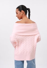 Sweater rosa off-the-shoulder com textura canelada e design de malha de cabos, apresentando um decote largo e mangas soltas, combinado com jeans azuis.