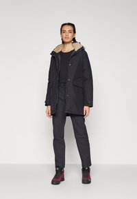 Veste noire imperméable avec une capuche en fourrure, poches avant et dos légèrement plus long. Associée à un pantalon noir et des bottes de randonnée avec des accents rouges.