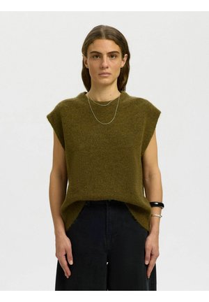 Femme aux cheveux bruns portant un pull sans manches vert olive, un pantalon noir, un collier en argent et des bracelets, debout devant un fond uni.