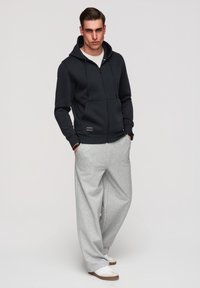 Jeune homme portant un sweat à capuche noir zippé, un pantalon de jogging large gris clair, des baskets blanches, debout les mains dans les poches.