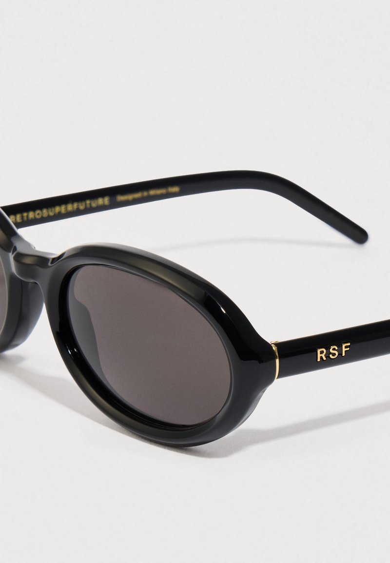 Lunettes de soleil ovales noires avec des verres sombres, monture en plastique brillante et accents dorés, arborant le logo "RSF" sur le côté.
